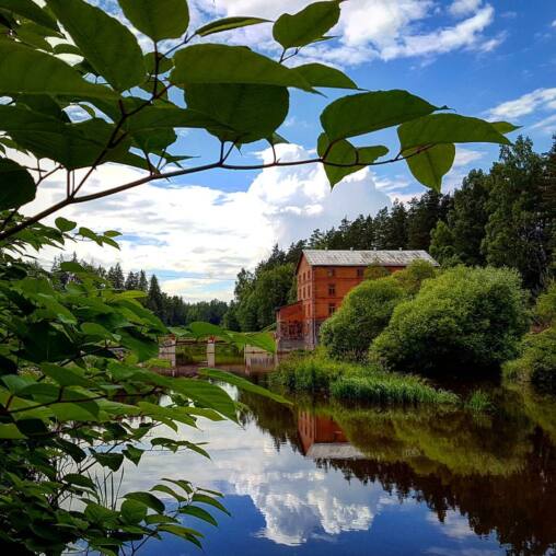 Red brick watermill beside a still river in summer, forest and clouds mirrored in the dark water, framed by overhanging leaves