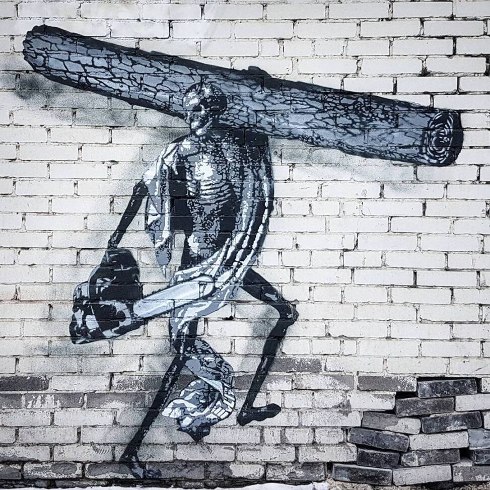Black and white stencil street art of a skeletal figure striding with a felled log across its shoulders and a chainsaw in hand, on a whitewashed brick wall