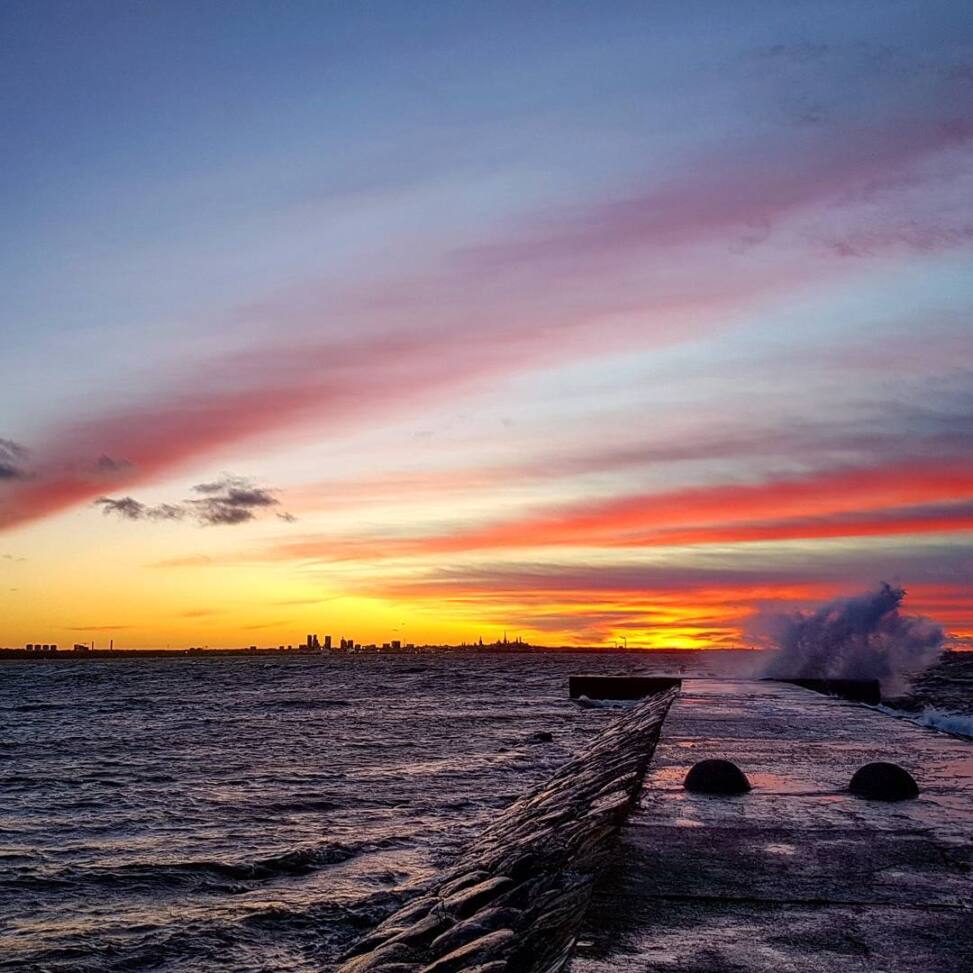 Streaked pink and orange sunset over the Tallinn skyline, waves breaking against a concrete pier in the foreground