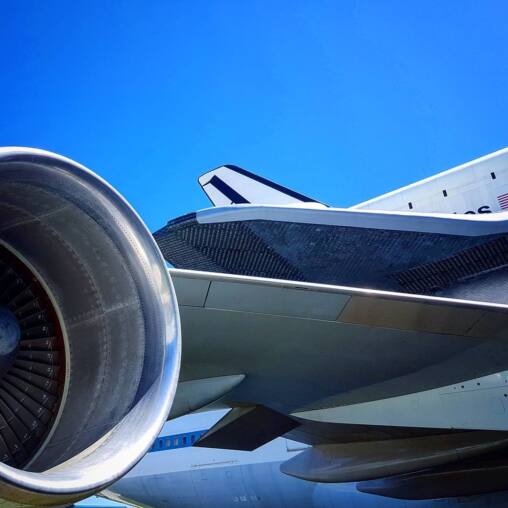 Jet engine nacelle in the foreground beneath the wing of a Boeing 747 carrying a space shuttle, against a cloudless blue sky
