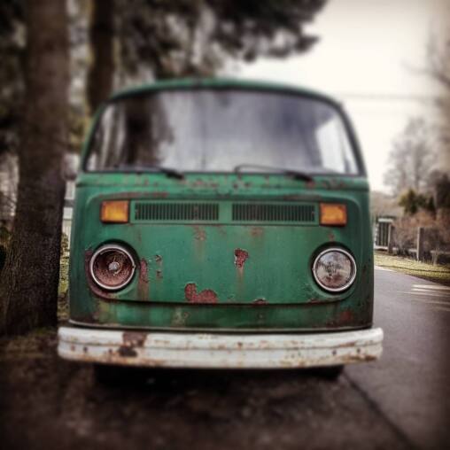 Front of a rusting green Volkswagen Type 2 van parked under bare trees, its chrome bumper pitted and paint flaking