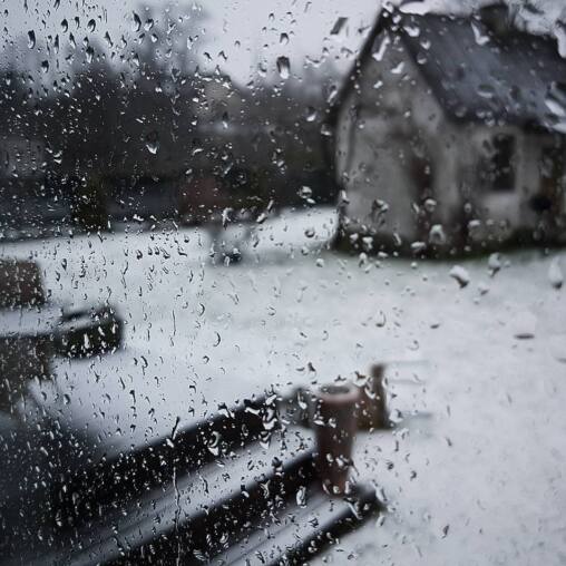 Fresh snow on a garden seen through a window streaming with raindrops, a white gabled house blurred beyond