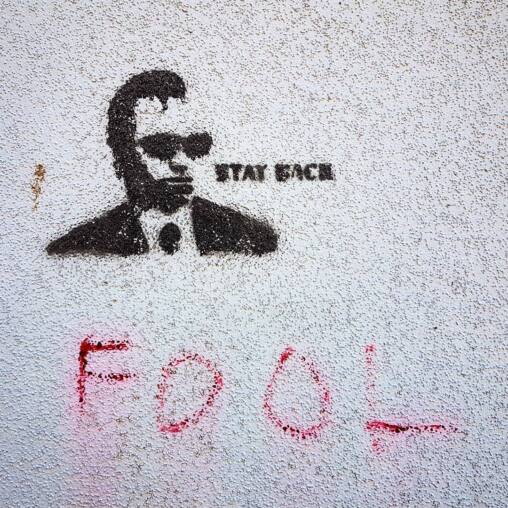 Stencil graffiti of a man in sunglasses and a suit captioned STAY BACK, with FOOL sprayed in pink beneath on a white wall