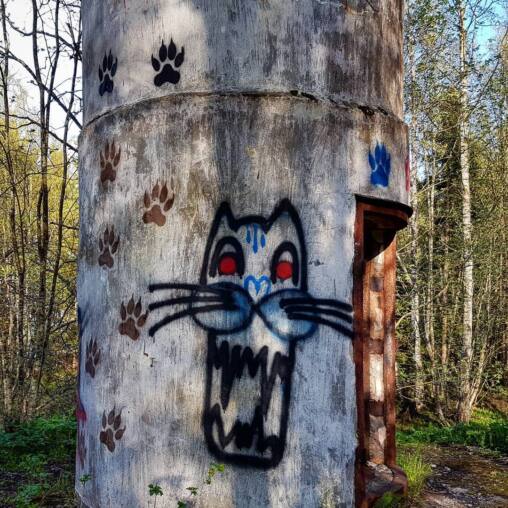 Grinning cat face with red eyes sprayed on a concrete water tower, painted paw prints climbing the wall around it