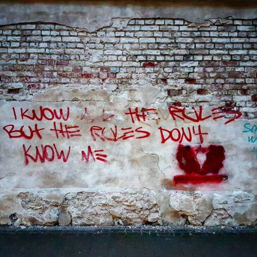 Red spray-painted words reading I know the rules but the rules don't know me on a crumbling plastered brick wall in Tartu
