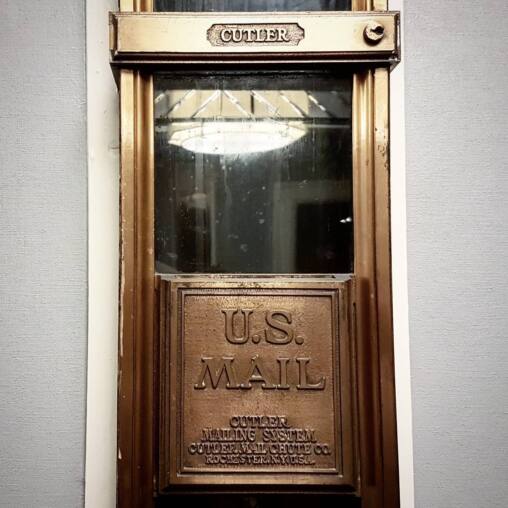 Brass US Mail chute door lettered Cutler Mailing System, Cutler Mail Chute Co, Rochester NY, set into a pale wall