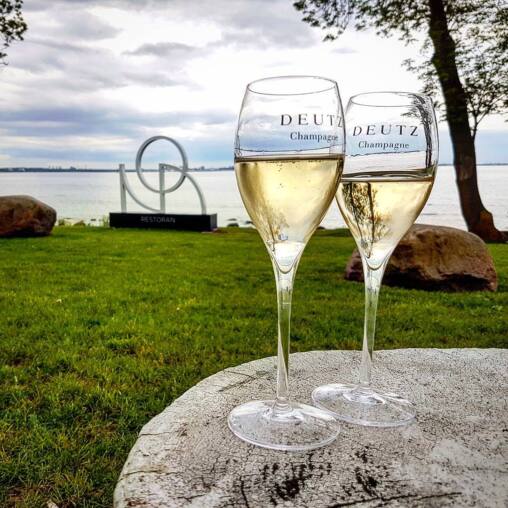 Two Deutz champagne flutes on a pale stone table on a lawn above the sea, the Noa restaurant sign and Tallinn skyline behind