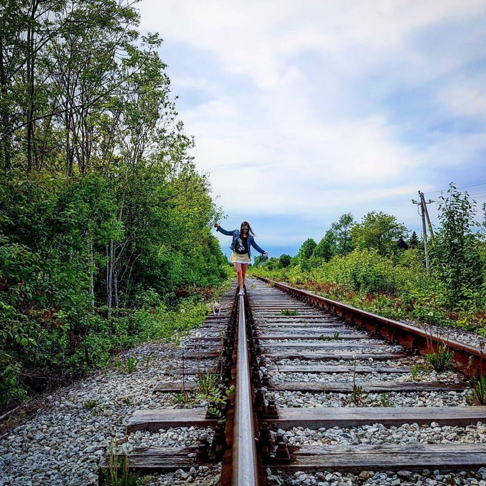 Woman in a denim jacket balancing along a disused railway rail with arms out, a chihuahua walking beside her between overgrown banks