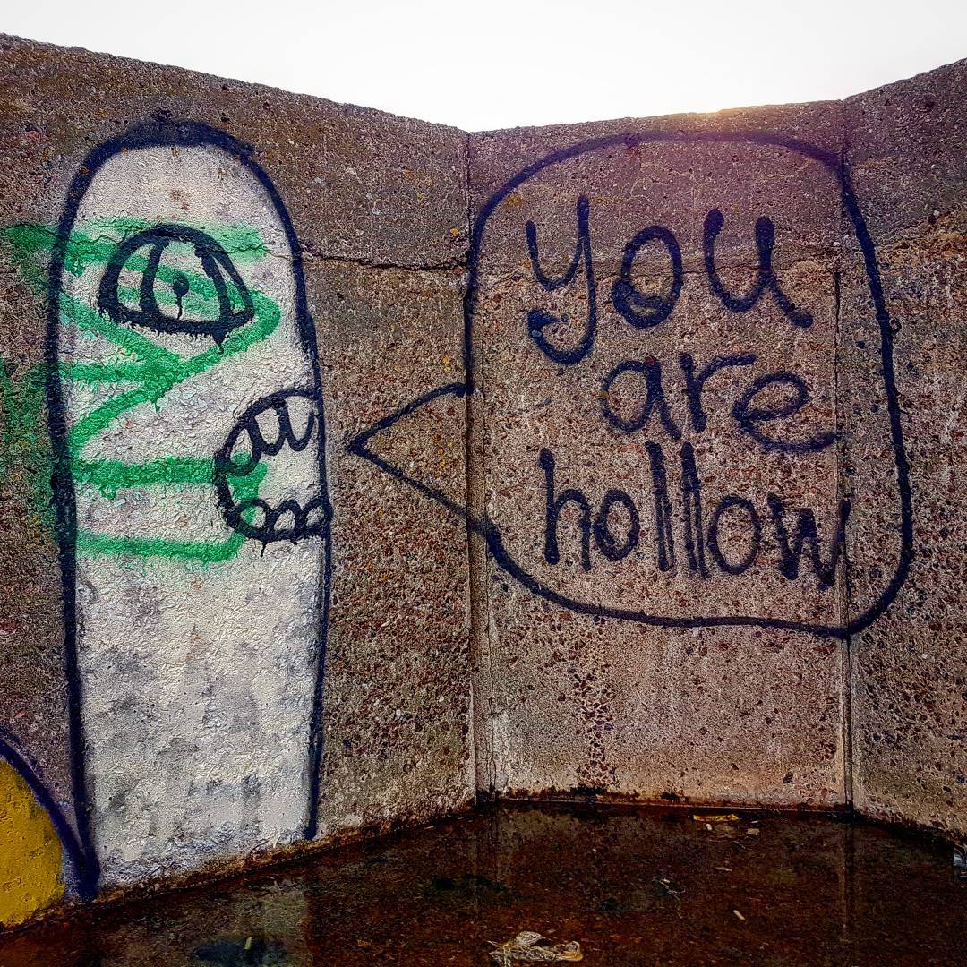 Graffiti of a pale toothy figure with one eye on a concrete corner, a speech bubble on the adjoining wall reading you are hollow