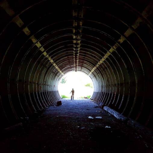 Silhouette of a person with a bicycle at the bright far end of a long corrugated steel tunnel, ribs curving away into darkness