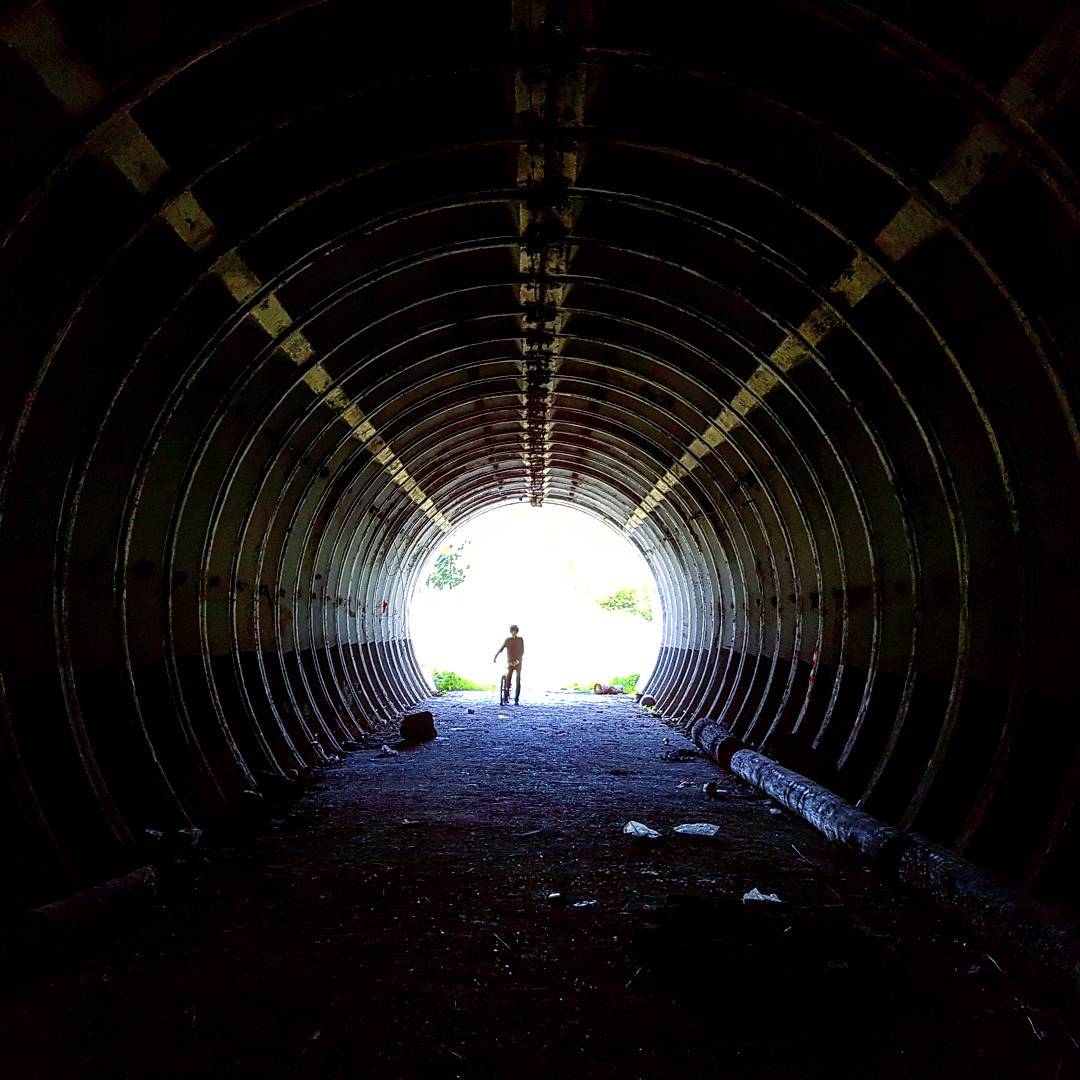 Silhouette of a person with a bicycle at the bright far end of a long corrugated steel tunnel, ribs curving away into darkness