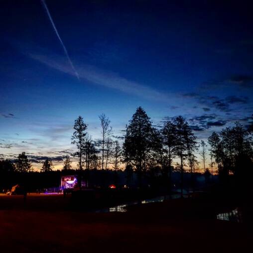Small lit concert stage glowing purple across a field at dusk, tall trees silhouetted against the last orange of sunset