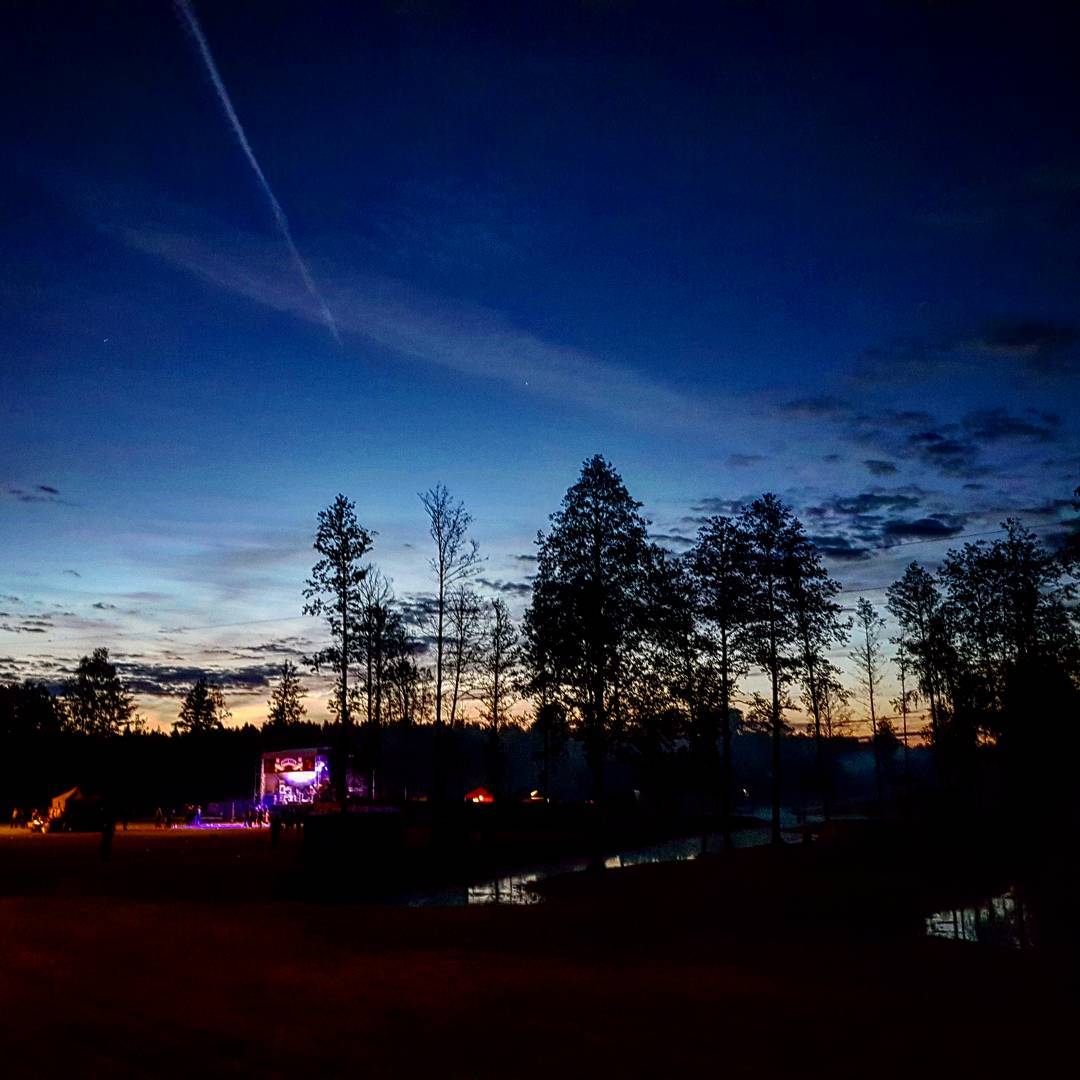 Small lit concert stage glowing purple across a field at dusk, tall trees silhouetted against the last orange of sunset