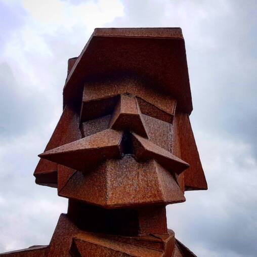Rusted Corten steel head sculpture with sharply faceted beard and hat, seen against heavy grey cloud