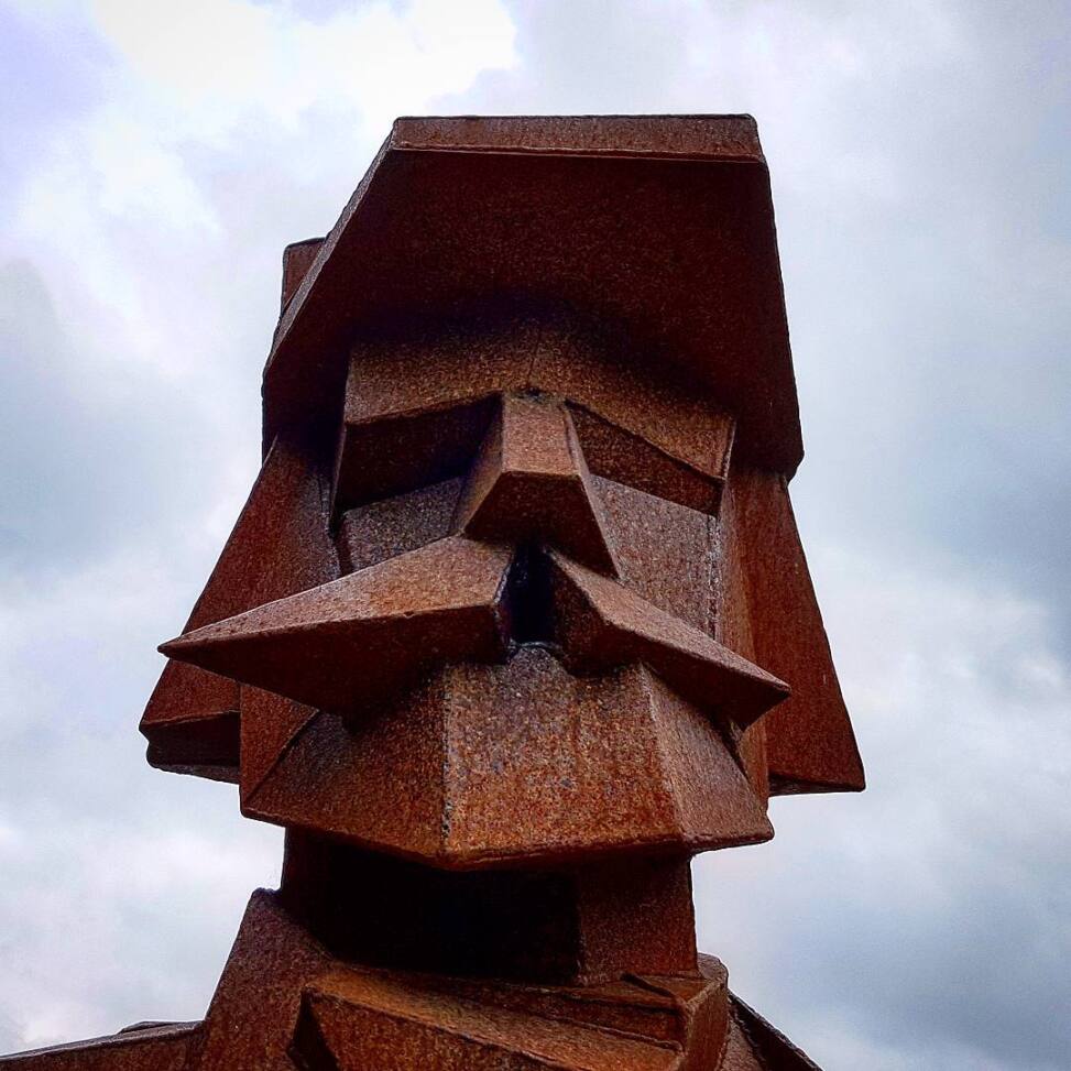 Rusted Corten steel head sculpture with sharply faceted beard and hat, seen against heavy grey cloud