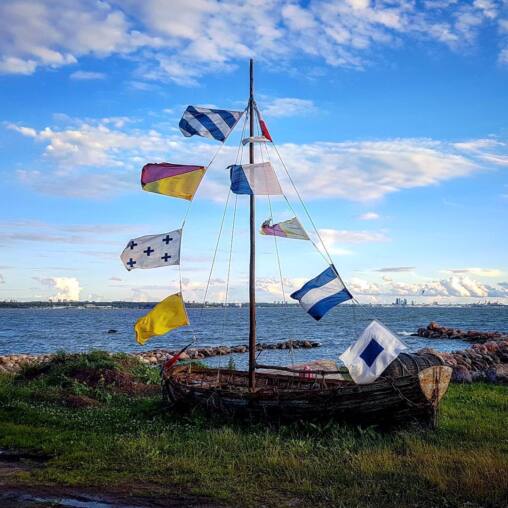 Old wooden boat set on the grass by the shore with a mast rigged full of coloured signal flags, the Tallinn skyline across the bay