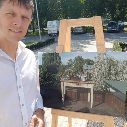 Man in a white shirt standing beside a large photograph mounted on a wooden easel outdoors, the print showing a laid table in a summer garden