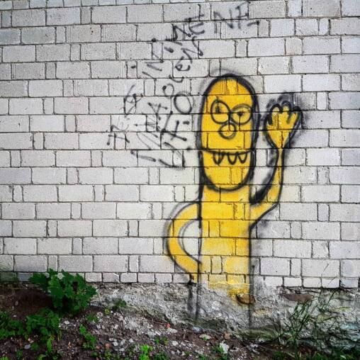 Yellow spray-painted grinning alien figure with a raised hand on a whitewashed brick wall, black tags above it