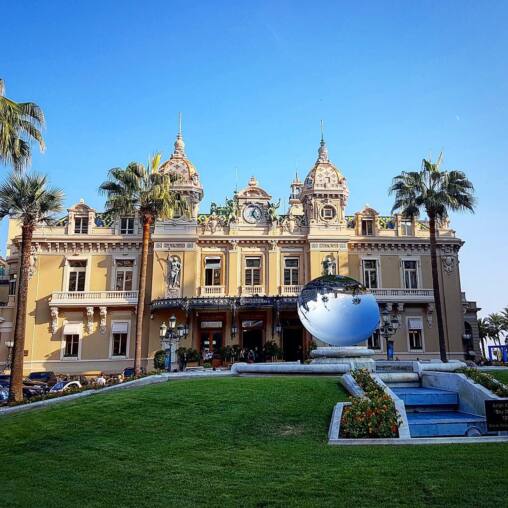 Belle Époque casino facade with twin domes and a clock, palms in front and a mirrored sphere sculpture on the lawn