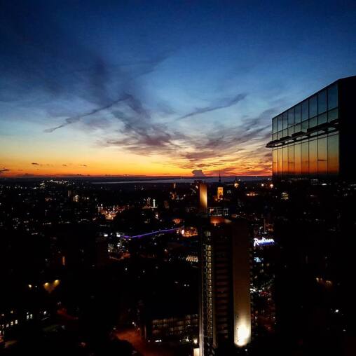 Dusk over Tallinn from a high floor, orange band of sunset along the horizon and a glass tower reflecting it, city lights below