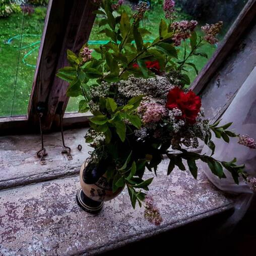 Bunch of spiraea, yarrow and a red carnation in a small vase on a peeling wooden window sill, the green garden beyond the glass