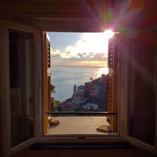 Open shuttered window framing the sea at sunset, a church campanile and ochre houses on the wooded slope below