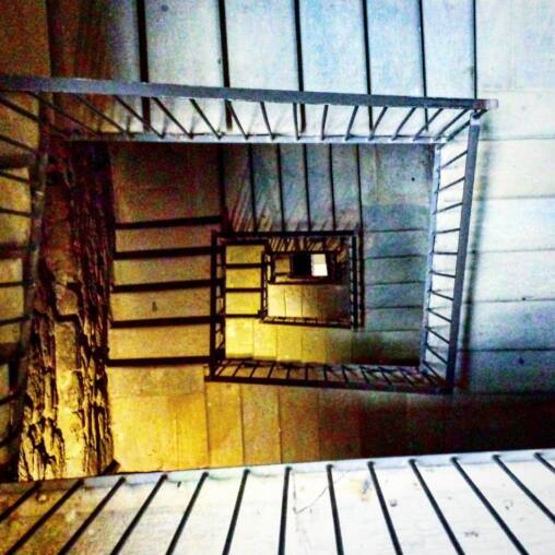Looking down a square stairwell with black metal railings, ochre walls and stone steps spiralling to a small bright square far below