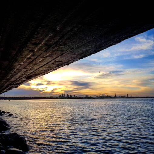 Sunset over the sea framed by the dark underside of a concrete pier, the Tallinn skyline in silhouette along the horizon
