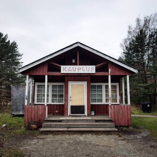 Red timber village shop with a hand-painted Kauplus sign over the porch, its door boarded over and windows blank