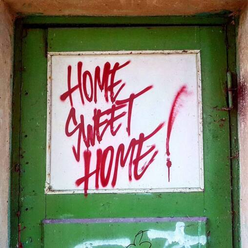 Red spray-painted words reading HOME SWEET HOME across the white panel of a peeling green door in a derelict building