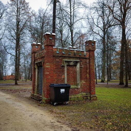Small red brick folly with crenellated turrets standing in a bare autumn park, a wheelie bin parked against its wall