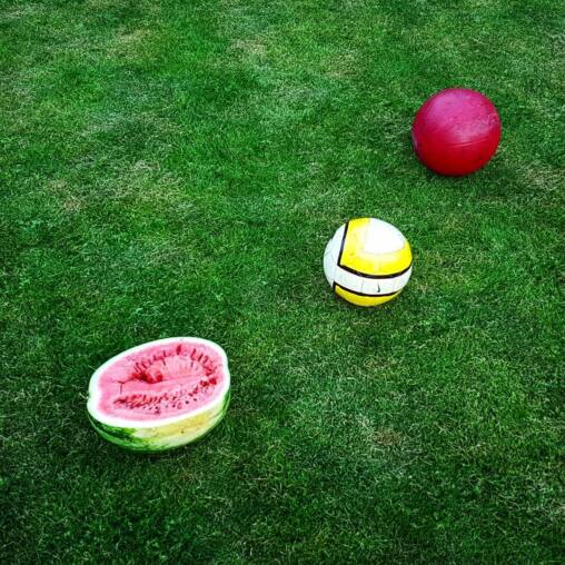 Half a watermelon, a yellow football and a red rubber ball spaced across bright mown grass, seen from above