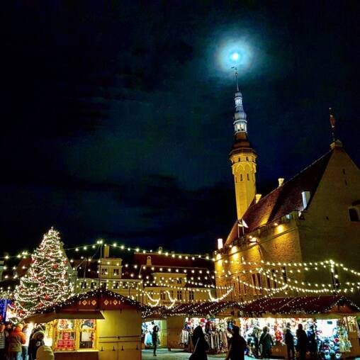 Halo-ringed moon above the town hall spire at night, the lit Christmas tree and market stalls strung with lights in the square below