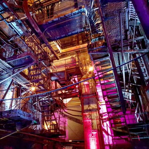 Looking up through the rusted steel gantries, ladders and pipework of the old power station, lit in magenta and gold