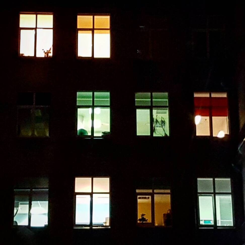 Grid of apartment windows lit at night in different colours, green, amber, red and white, the facade between them black