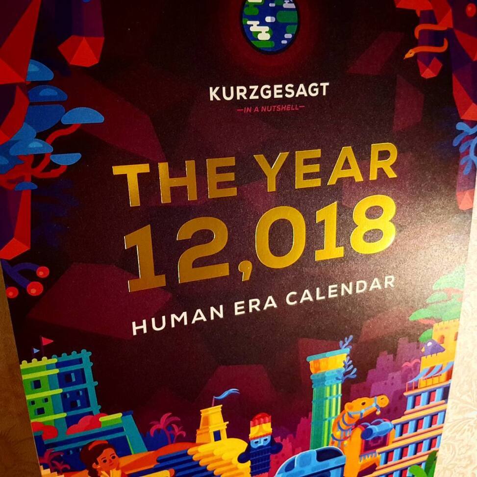 Kurzgesagt Human Era calendar cover in gold foil on deep red, illustrated with brightly coloured ancient cities and creatures